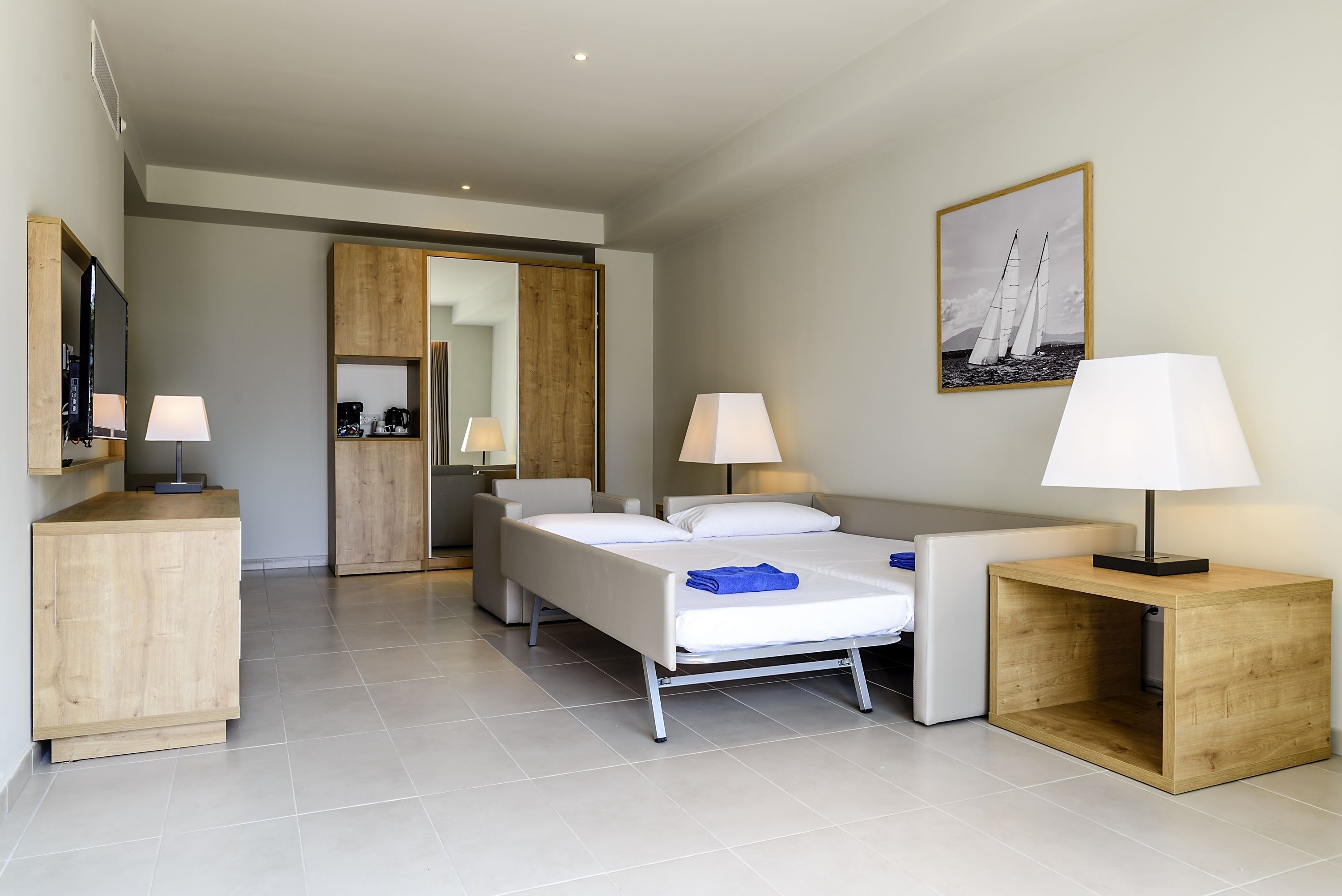 A modern hotel room with a bed, two bedside tables with lamps, a wooden nightstand, a TV, and a painting of sailboats.