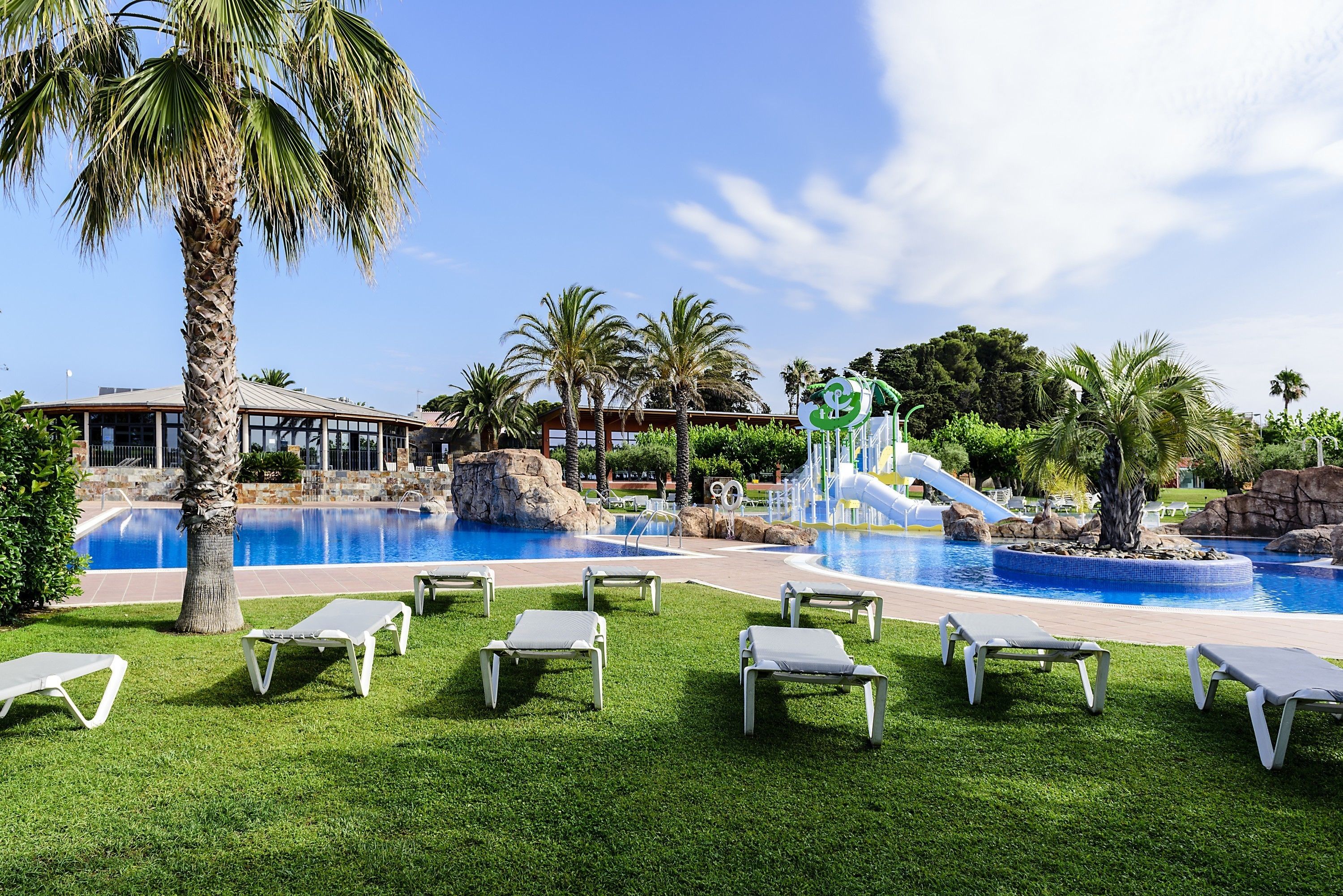 A pool area with multiple lounge chairs, a water slide, and palm trees.