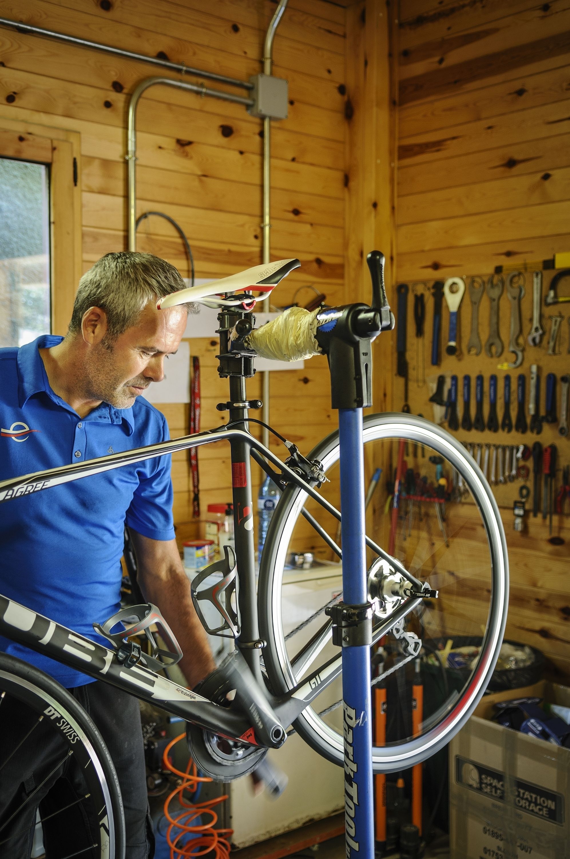 A man working on a bicycle in a workshop with wooden walls and tools hanging on the wall.