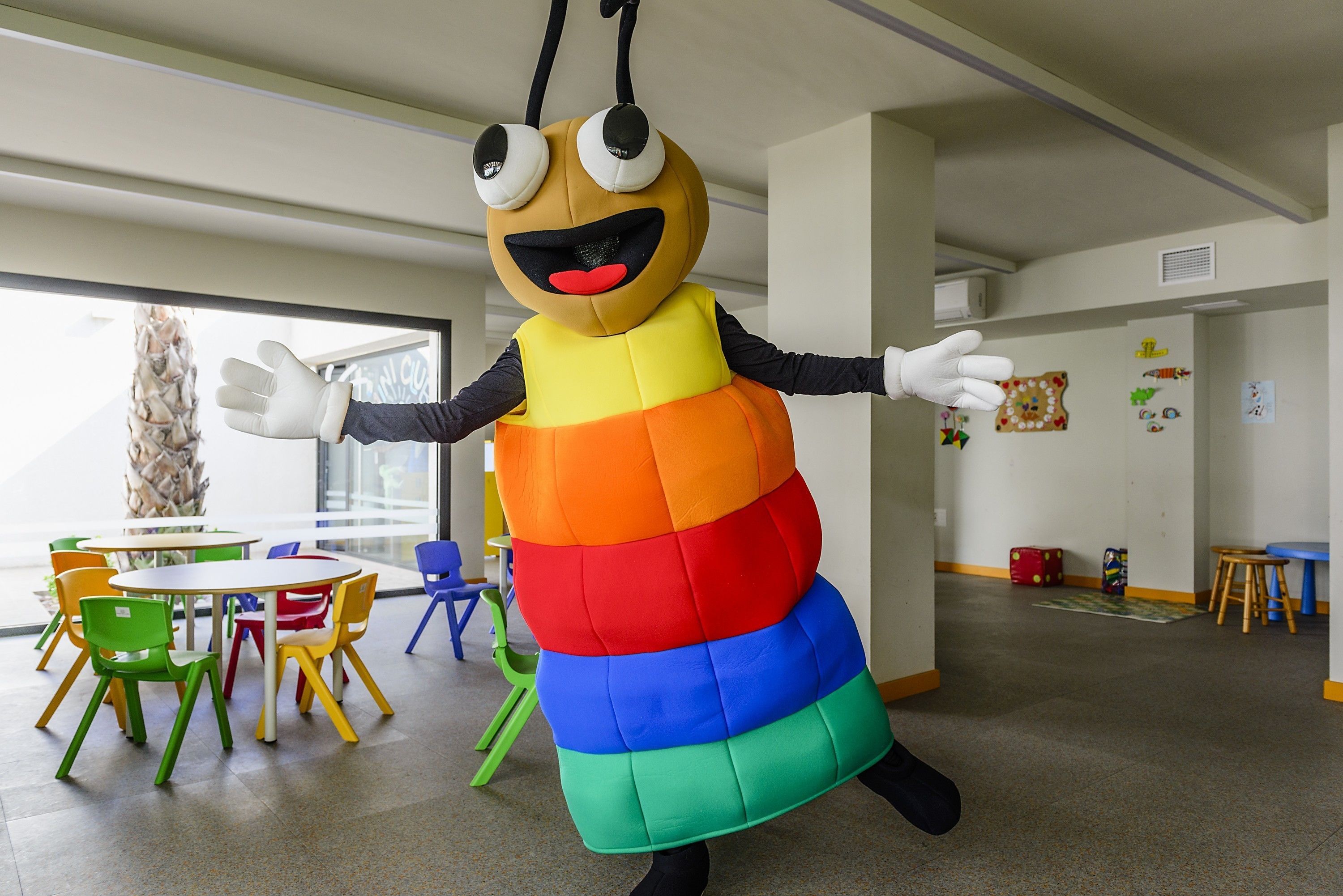 Una persona con un disfraz colorido en una habitación con un ventanal grande, una mesa con sillas y una pared con dibujos infantiles.