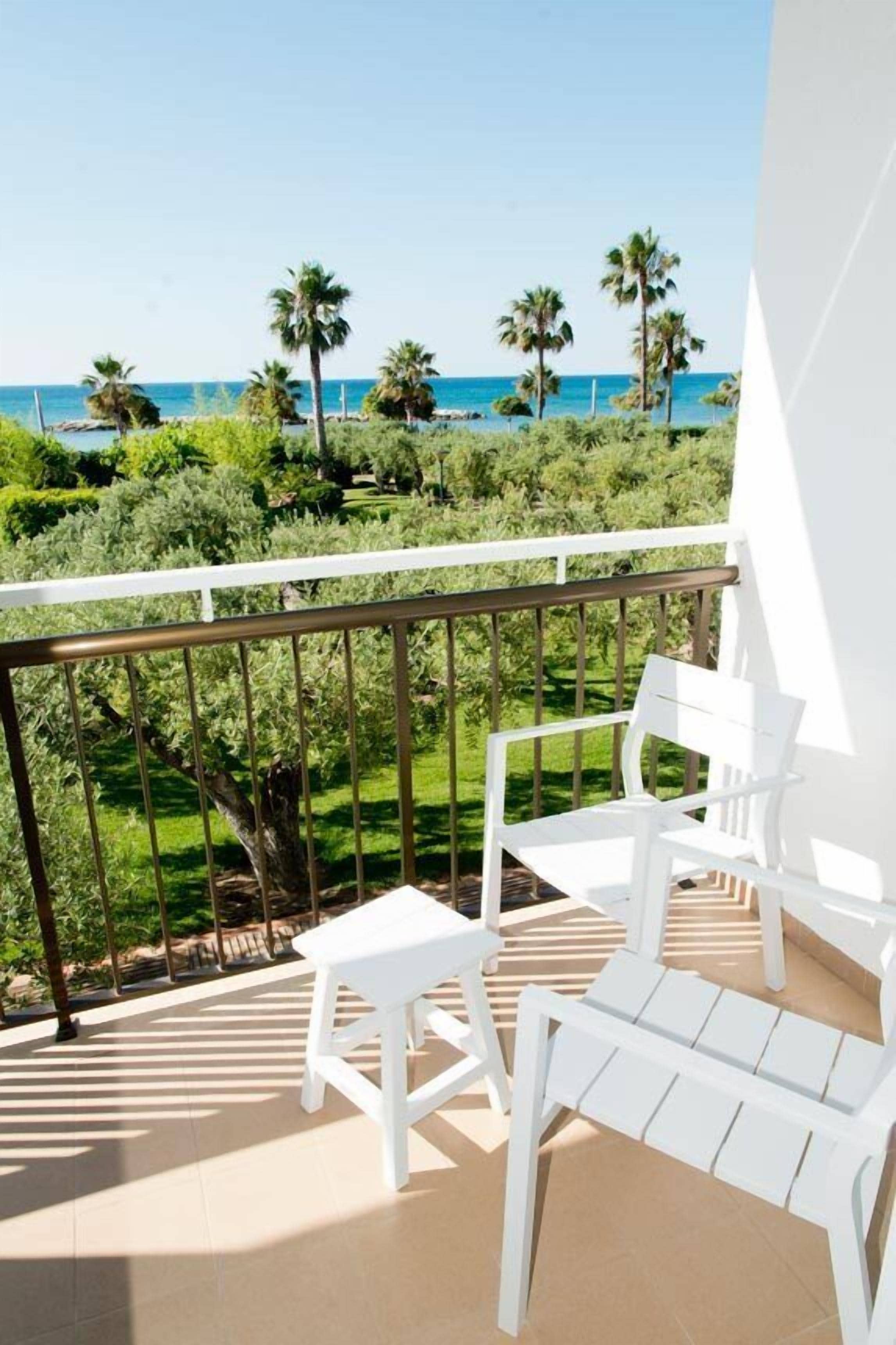 Un balcón con sillas blancas y una pequeña mesa, con vista a un jardín de palmeras y al mar.