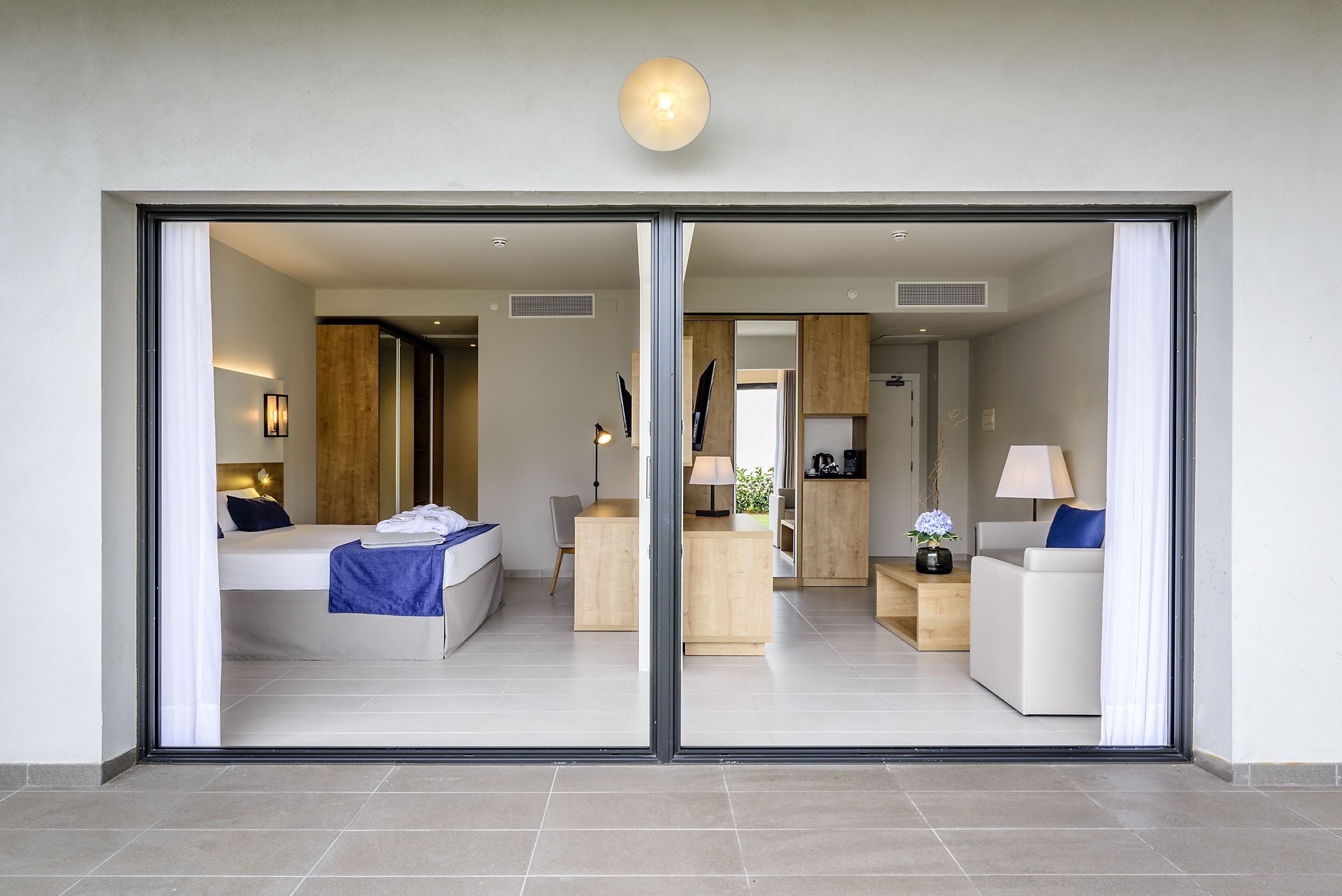 Ein modernes Hotelzimmer mit einer großen Glasschiebetür, einem Bett mit blauer Decke, einem Schreibtisch mit Lampe und einer Sitzecke mit weißem Sofa und blauem Kissen.