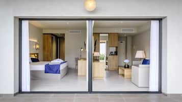 A modern hotel room with a large sliding glass door, a bed with a blue blanket, a desk with a lamp, and a seating area with a white sofa and a blue pillow.