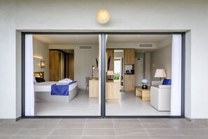 A modern hotel room with a large sliding glass door, a bed with a blue blanket, a desk with a lamp, and a seating area with a white sofa and a blue pillow. - Estival Eldorado (Cambrils)