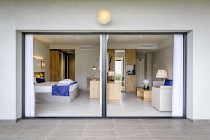 Una habitación de hotel moderna con un amplio ventanal corredera, una cama con una manta azul, un escritorio con una lámpara y una zona de estar con un sofá blanco y una almohada azul.