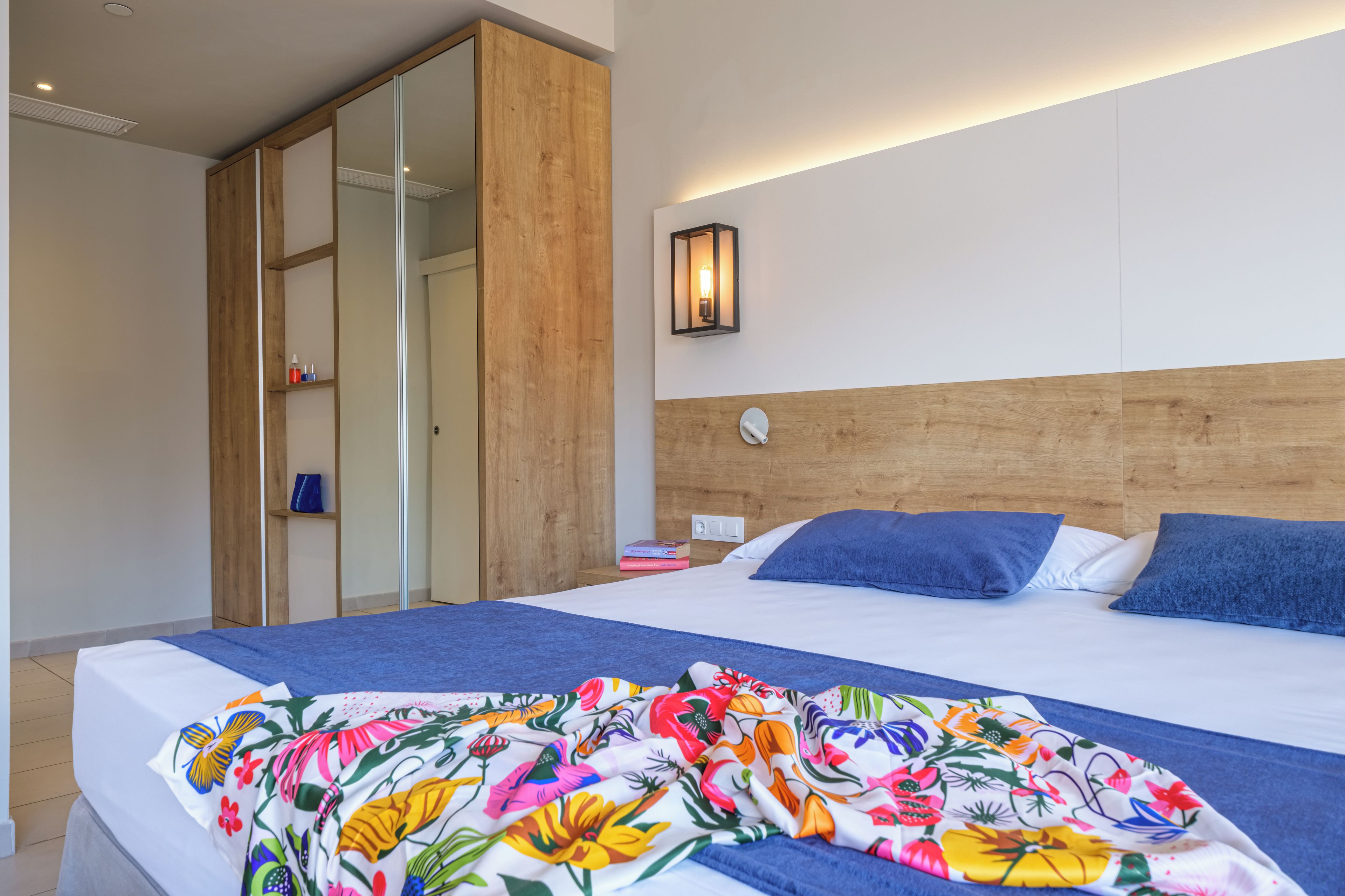 A modern hotel room with a wooden headboard, a large bed with a colorful floral bedspread, a nightstand with a lamp, and a sliding door closet.