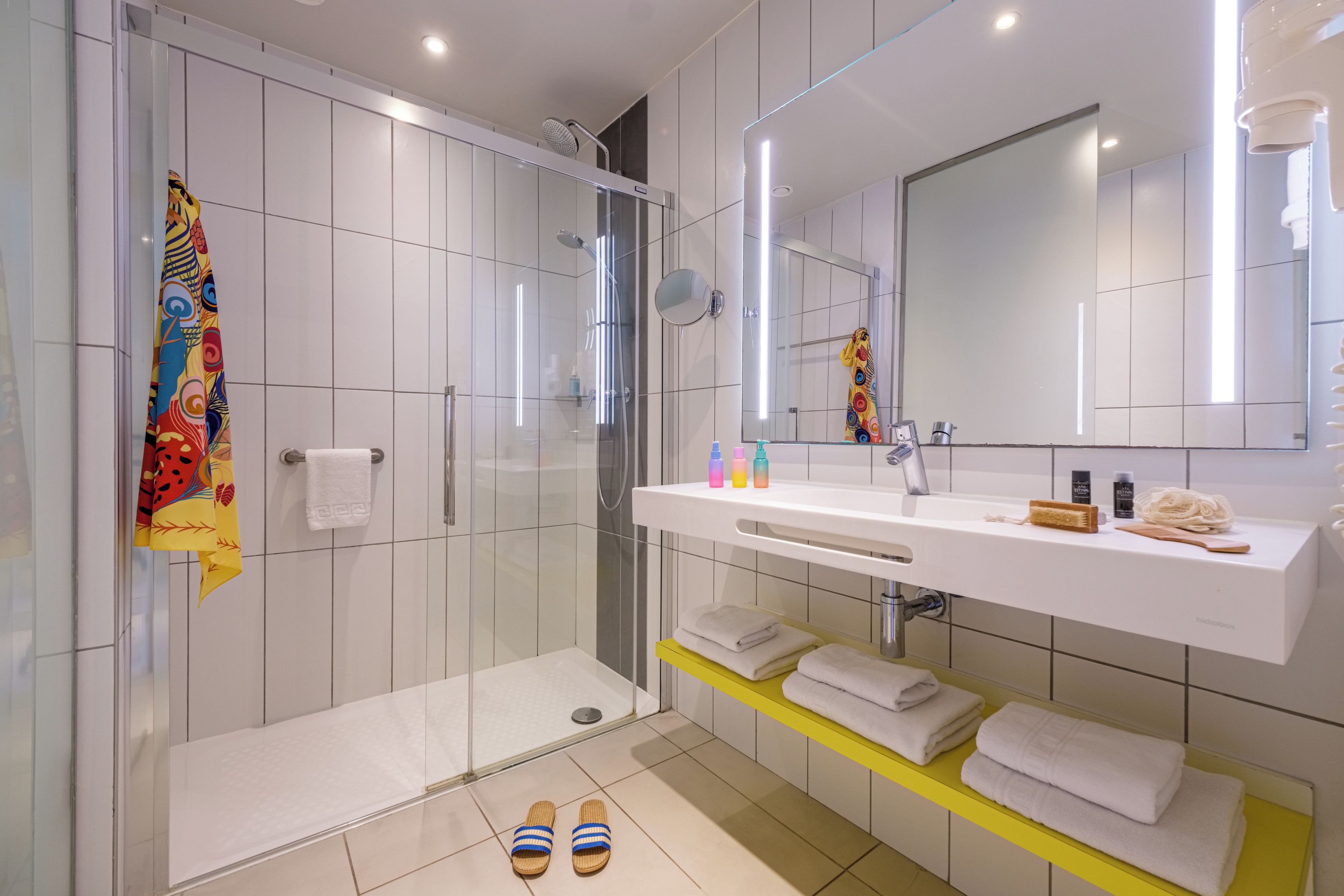 Een moderne badkamer met een douchecabine van glas, een witte wastafel met spiegel erboven en netjes opgestelde handdoeken op planken.