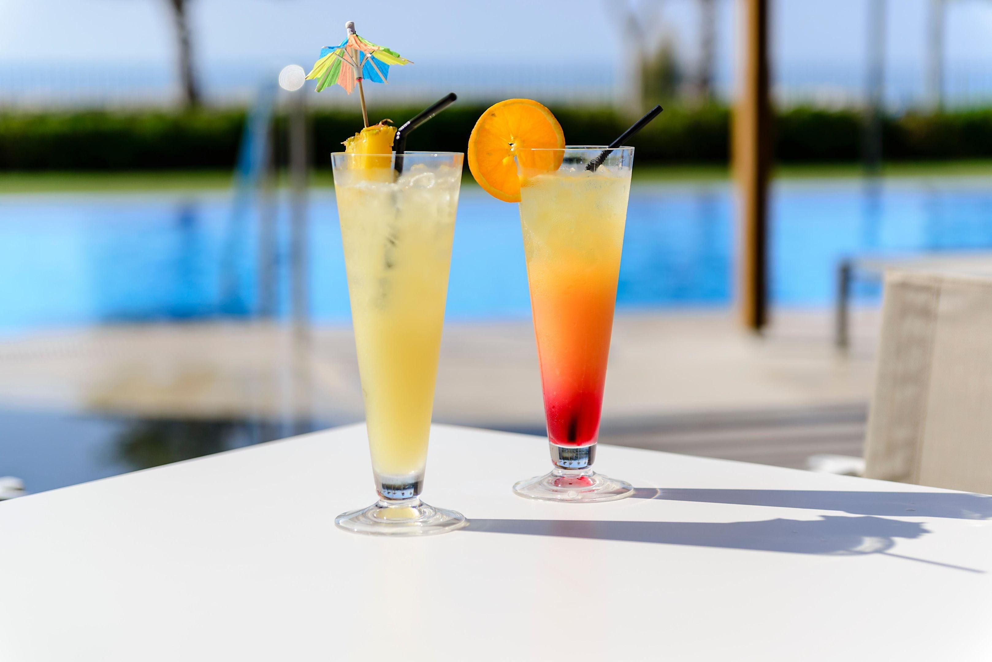 Two cocktails on a table by a pool with a clear view of the water and greenery in the background.