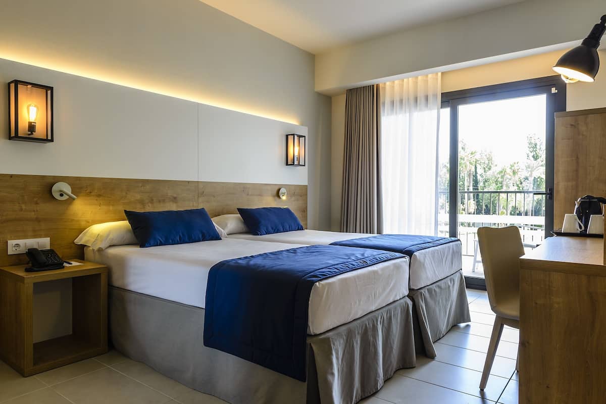 A hotel room with two beds, a desk, a chair, a phone on the nightstand, and a view of the outdoors.