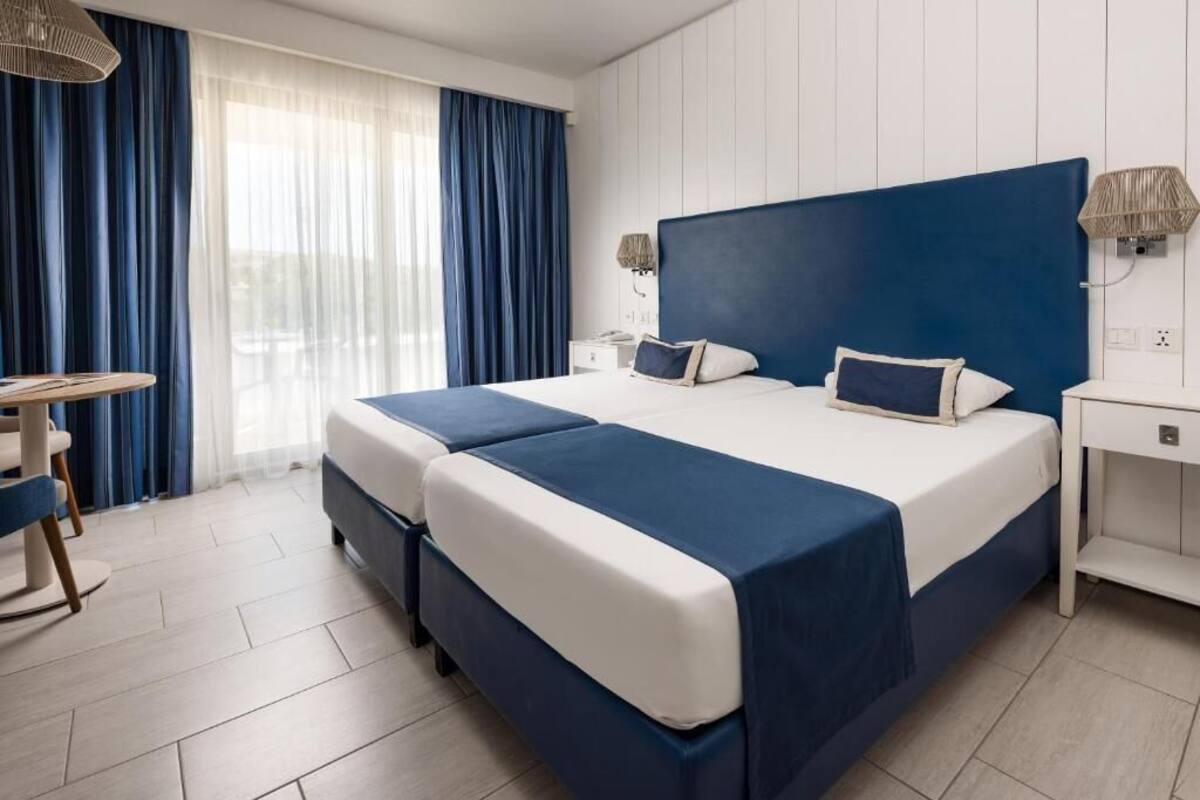 A double bed with a blue headboard, two bedside tables, a lamp, and a view of the outdoors through sheer curtains.