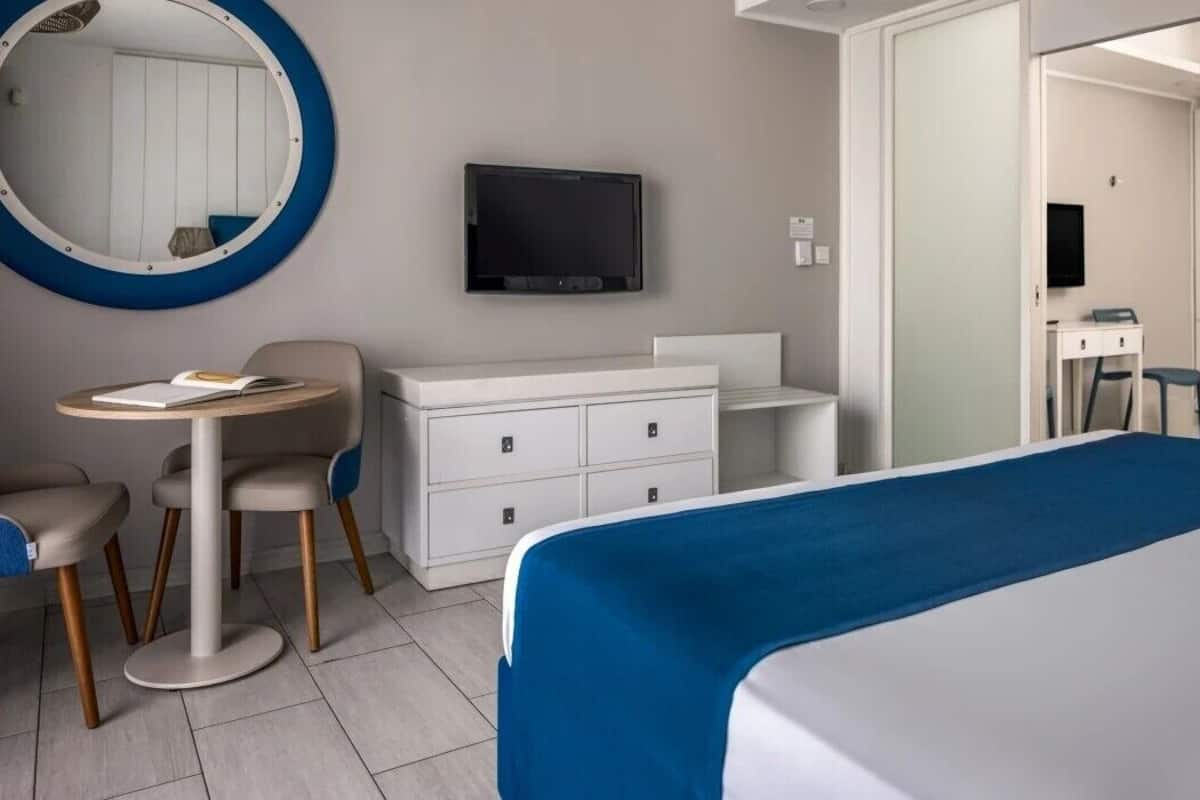 A hotel room with a bed, a desk with a chair, a round table with two chairs, a TV mounted on the wall, and a wardrobe.