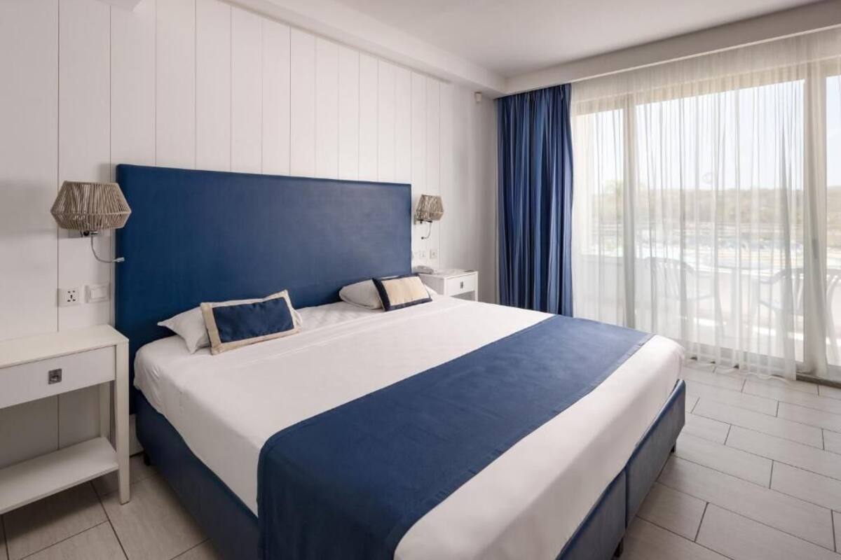A hotel room with a large bed, white walls, and a view of the outdoors.