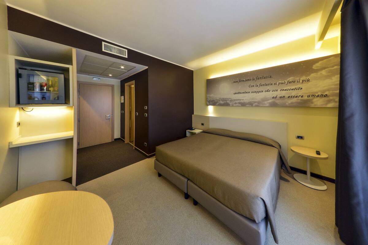 A hotel room with a bed, bedside tables, a desk with a computer, a small round table, and a wall-mounted artwork.