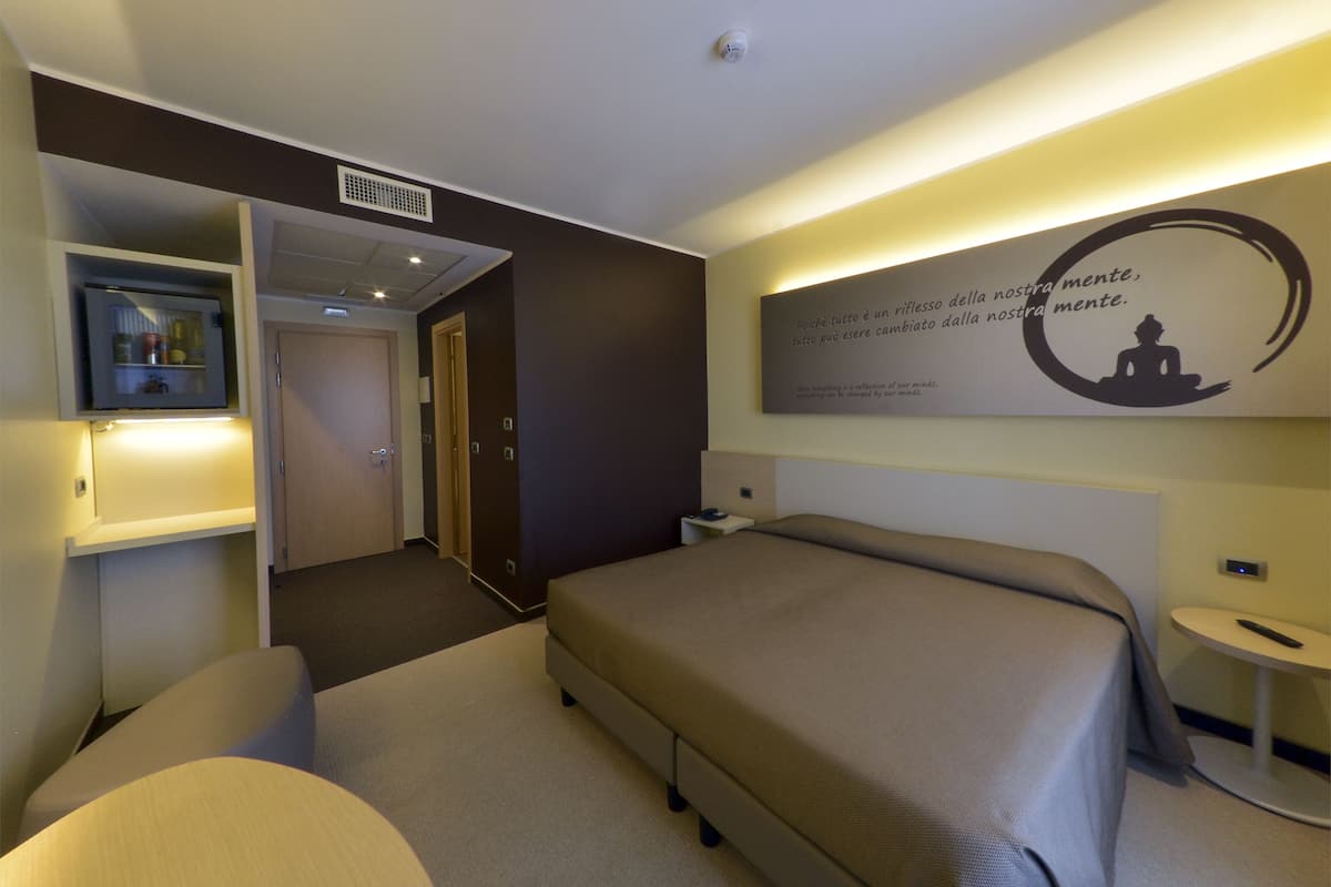 A hotel room with a bed, a desk, a chair, a small table, and a wall-mounted artwork.
