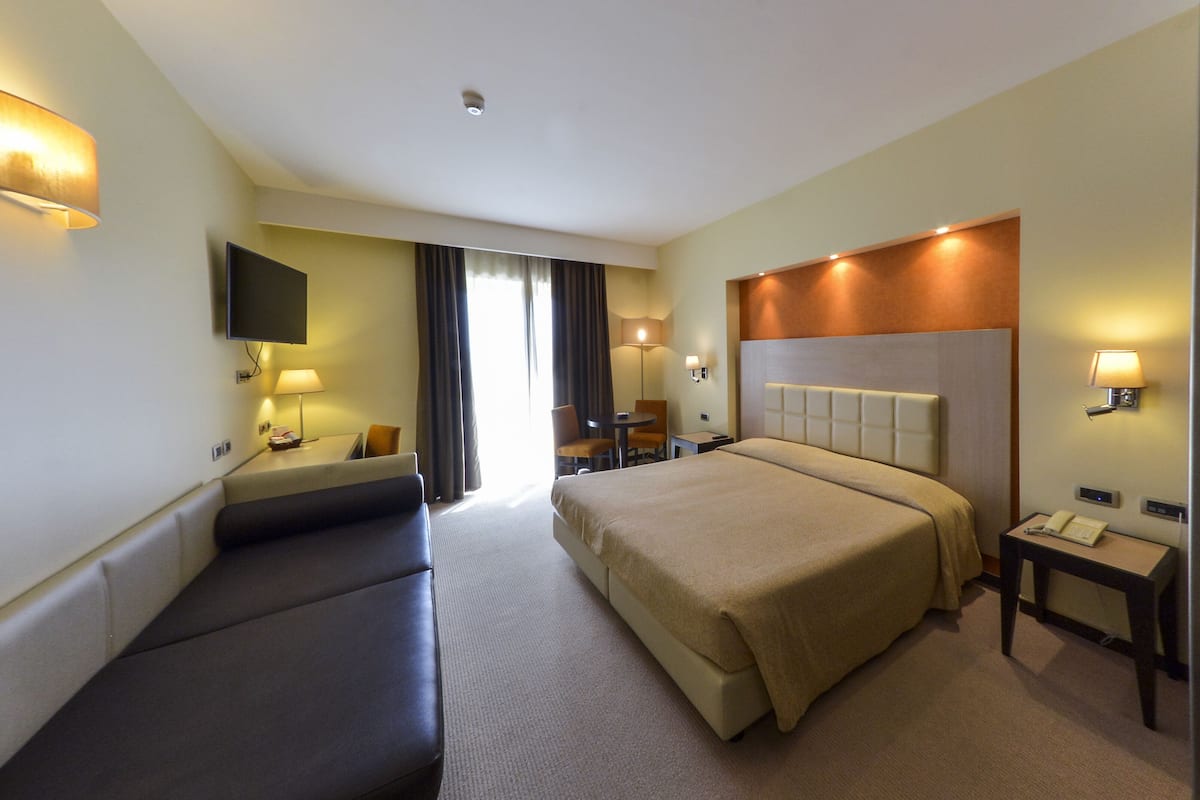 A hotel room with a large bed, a desk with a chair, a TV mounted on the wall, and a window with curtains.