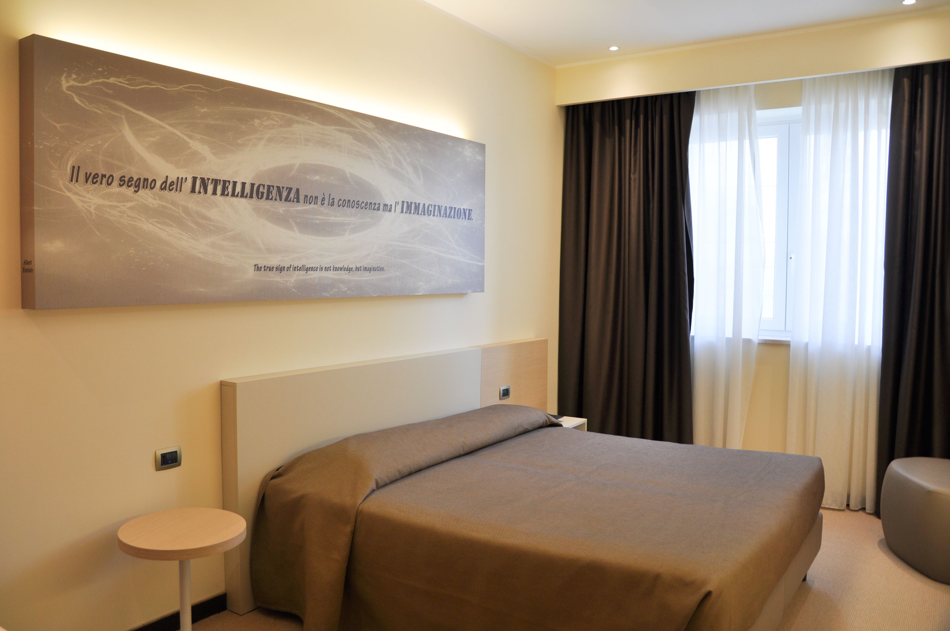 A hotel room with a large bed, a bedside table, a wall-mounted artwork, and a window with curtains.