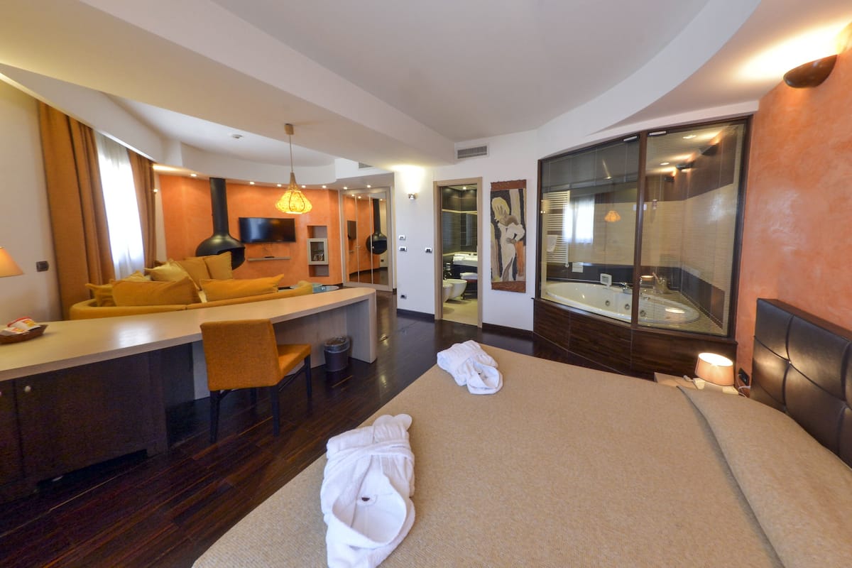 A modern hotel room with a large bed, a sitting area with a sofa, a dining table, and a large mirror.