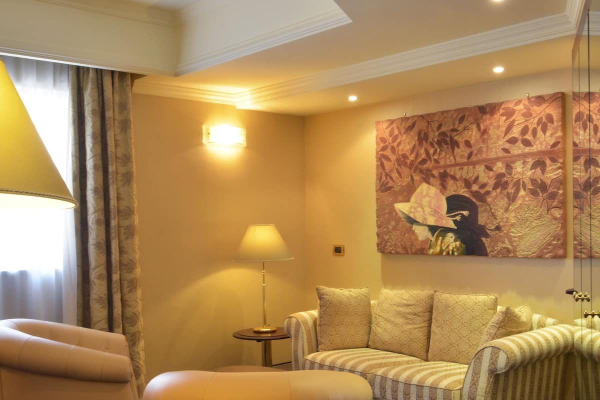 A hotel room with a sofa, armchair, and ottoman. There is a lamp, a painting on the wall, and a window with curtains.