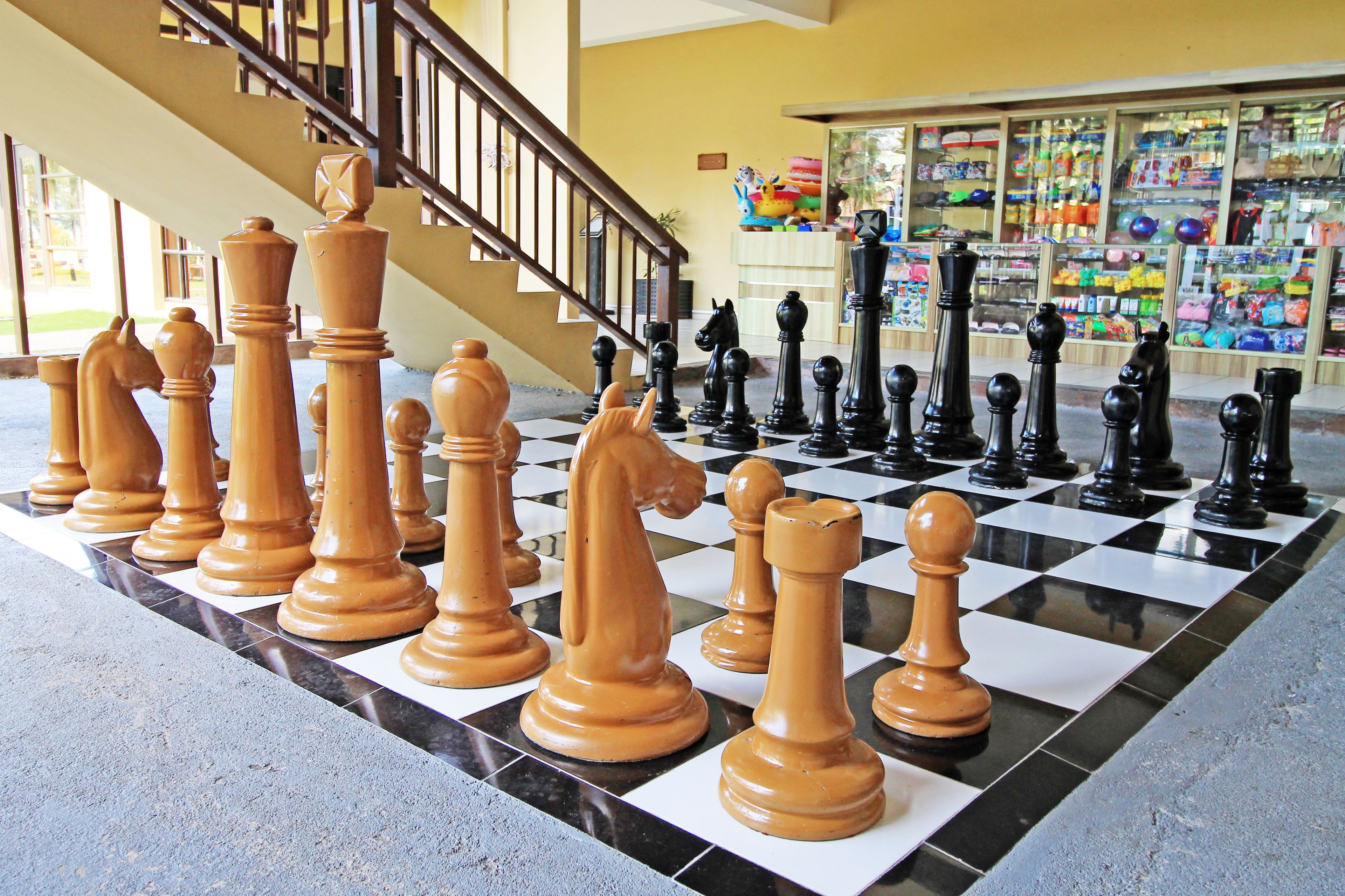 A large chessboard with oversized chess pieces in a lobby area with a staircase and a shop in the background.