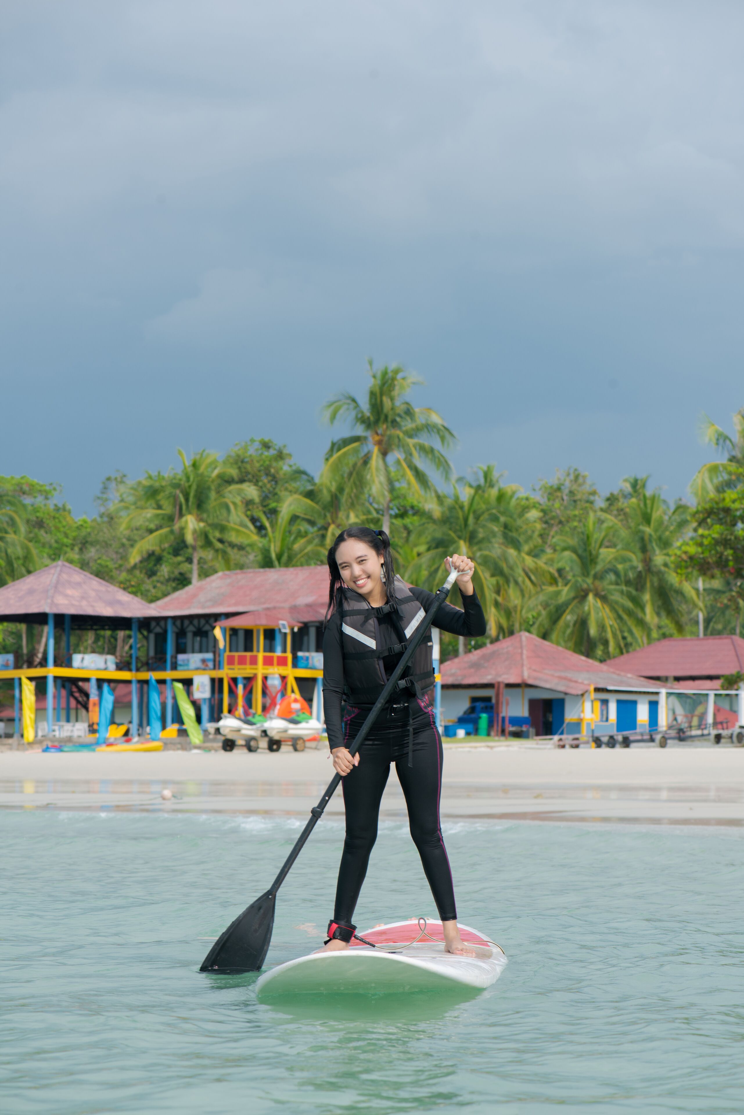 A person paddleboarding on calm water with a beach and palm trees in the background.