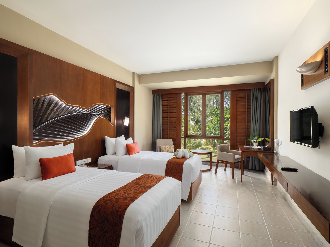 A hotel room with two beds, a desk, a chair, a TV, and large windows with a view of greenery.