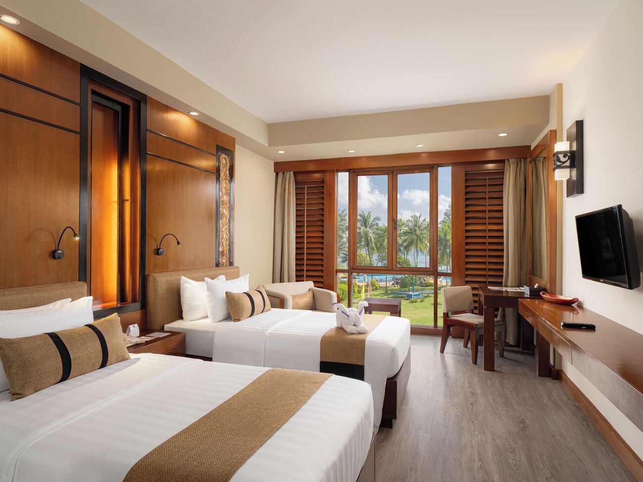 A hotel room with two beds, a desk, a TV, and large windows with a view of palm trees.