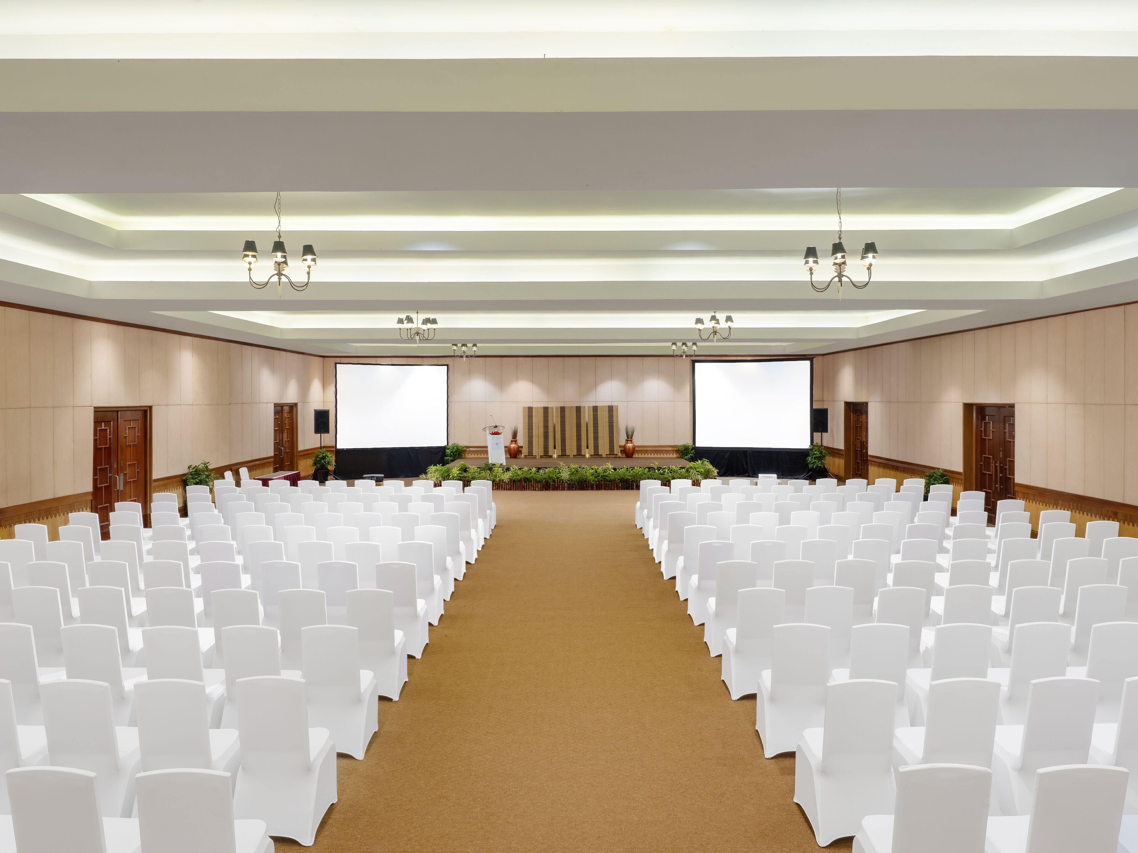 Un salón de conferencias con filas de sillas blancas, un escenario con una pantalla grande y candelabros.