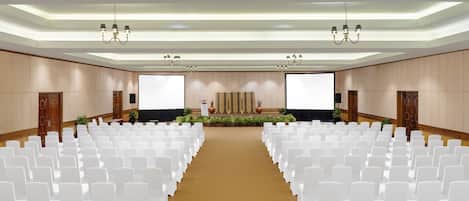 A conference room with rows of white chairs, a stage with a large screen, and chandeliers.