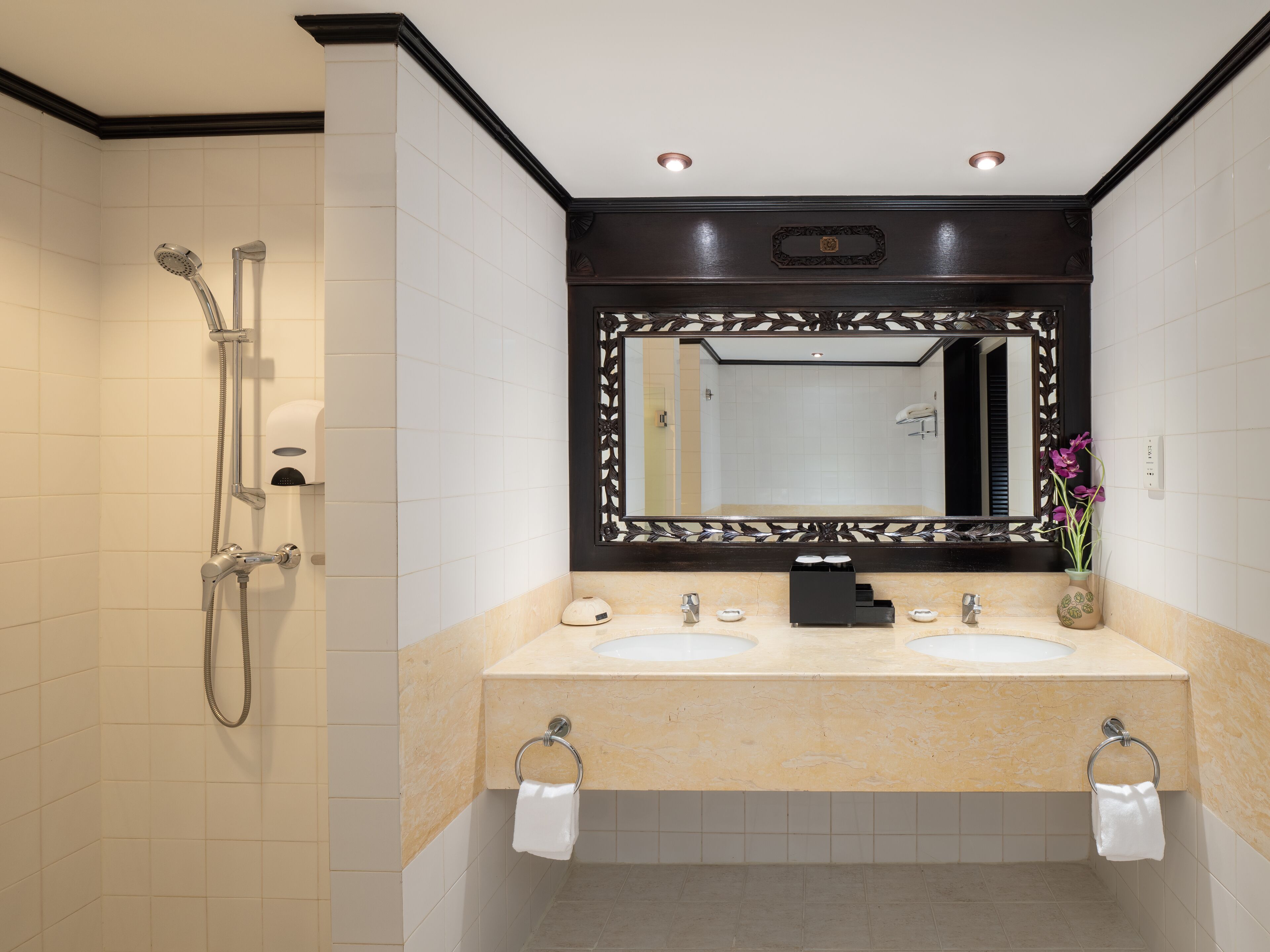Un baño moderno con tocador doble lavamanos, un espejo grande con marco decorativo, ducha y un florero con flores.