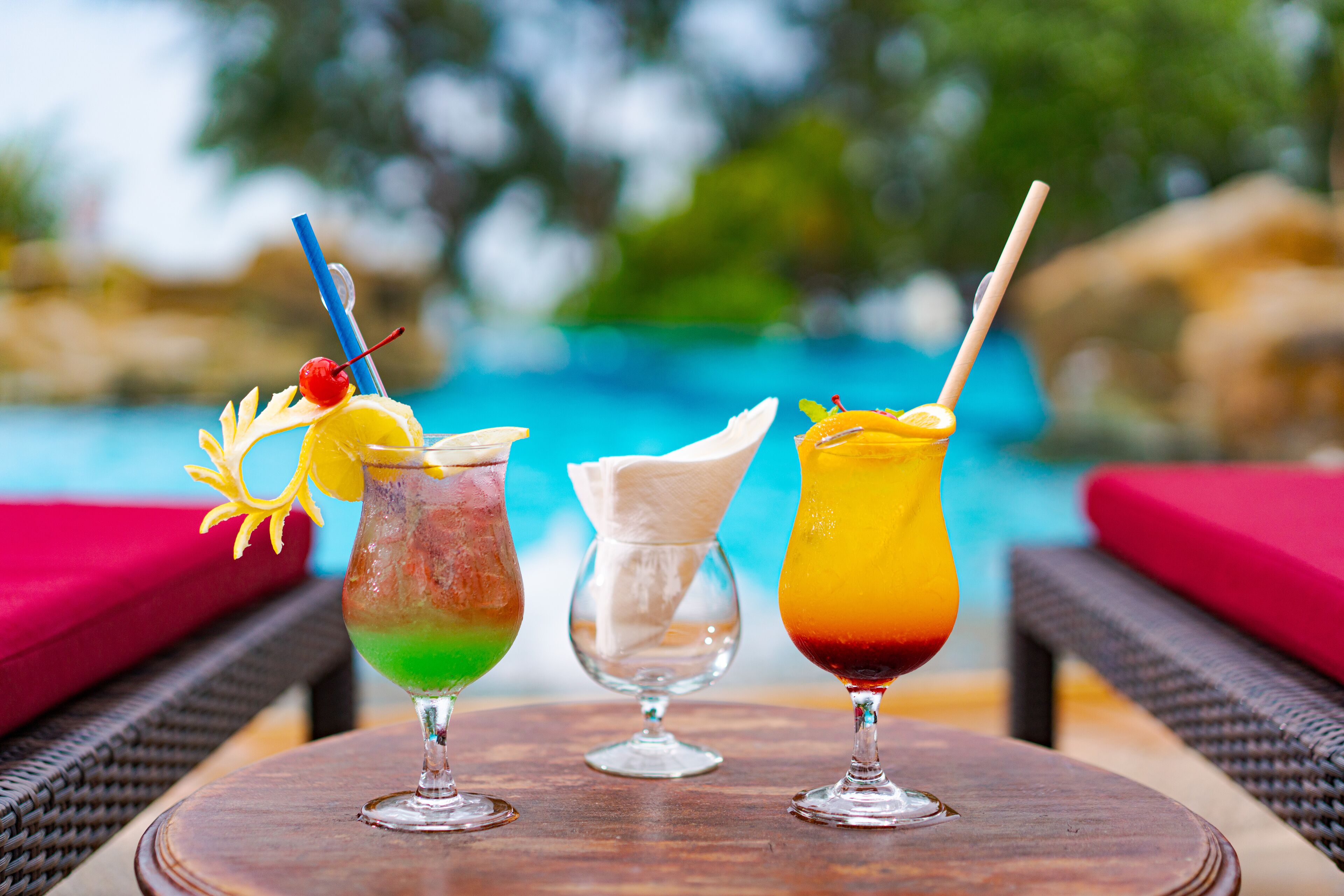 Three colorful cocktails on a wooden table by a poolside with wicker chairs and red cushions.