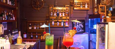 A bar area with two drinks on the counter, a ship's wheel on the wall, and shelves with bottles.