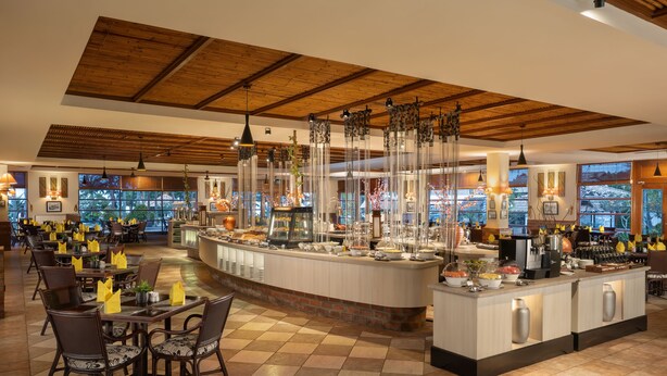 A spacious restaurant with a high ceiling and a central bar area. Tables are arranged around the bar, and the floor has a patterned tile design.