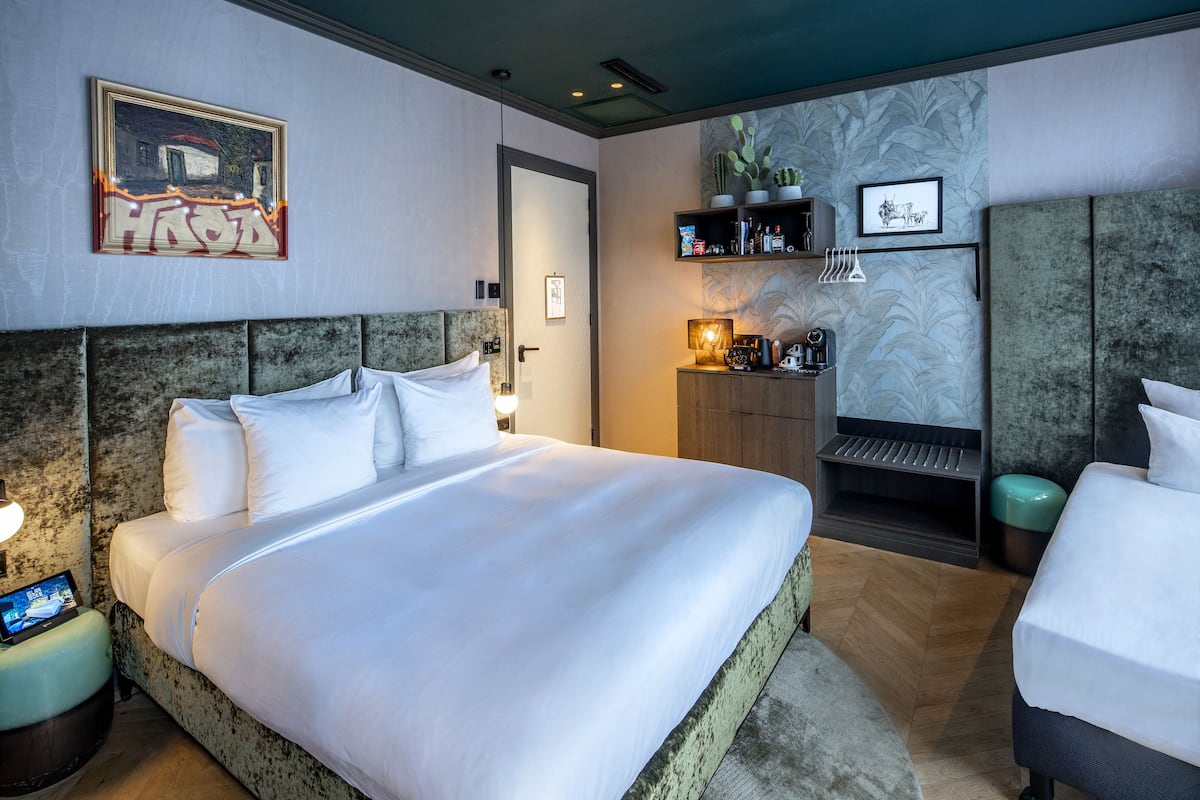 Camera d'albergo con un letto grande, un comodino con una lampada, un piccolo angolo salotto con una panca e una parete con una mensola a forma di cactus.