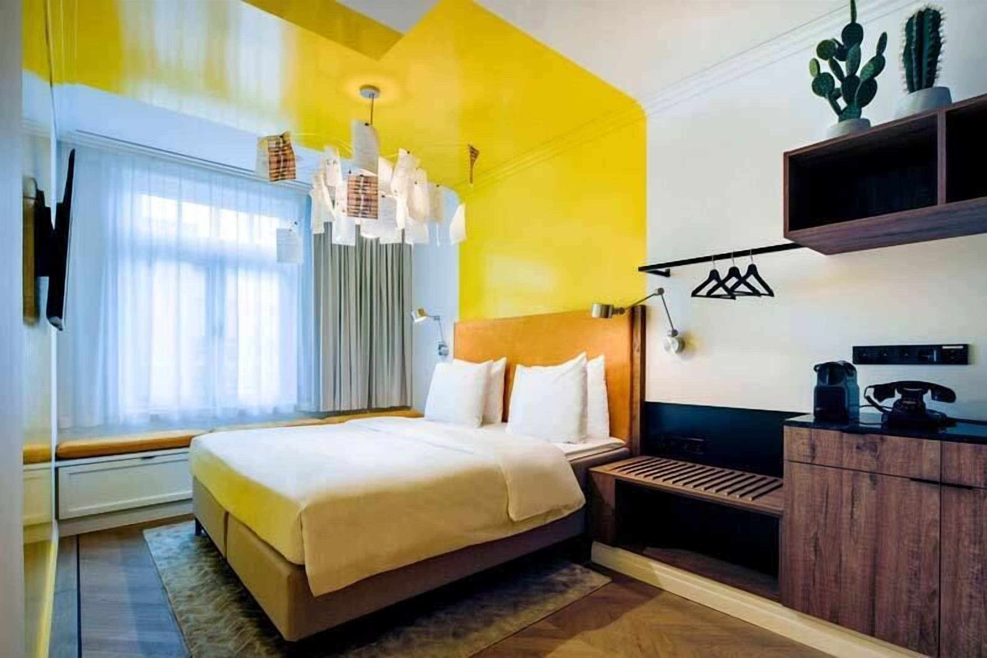 Una habitación de hotel moderna con una cama grande, una mesita de noche, un escritorio con una silla, un armario y una ventana con cortinas.