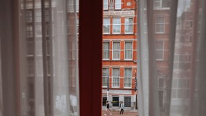 A view through a window with curtains, revealing a red brick building with a sign that reads "KASTEEL VAN AEMSTEL" and a street scene with pedestrians. - The Highlander (Amsterdam)