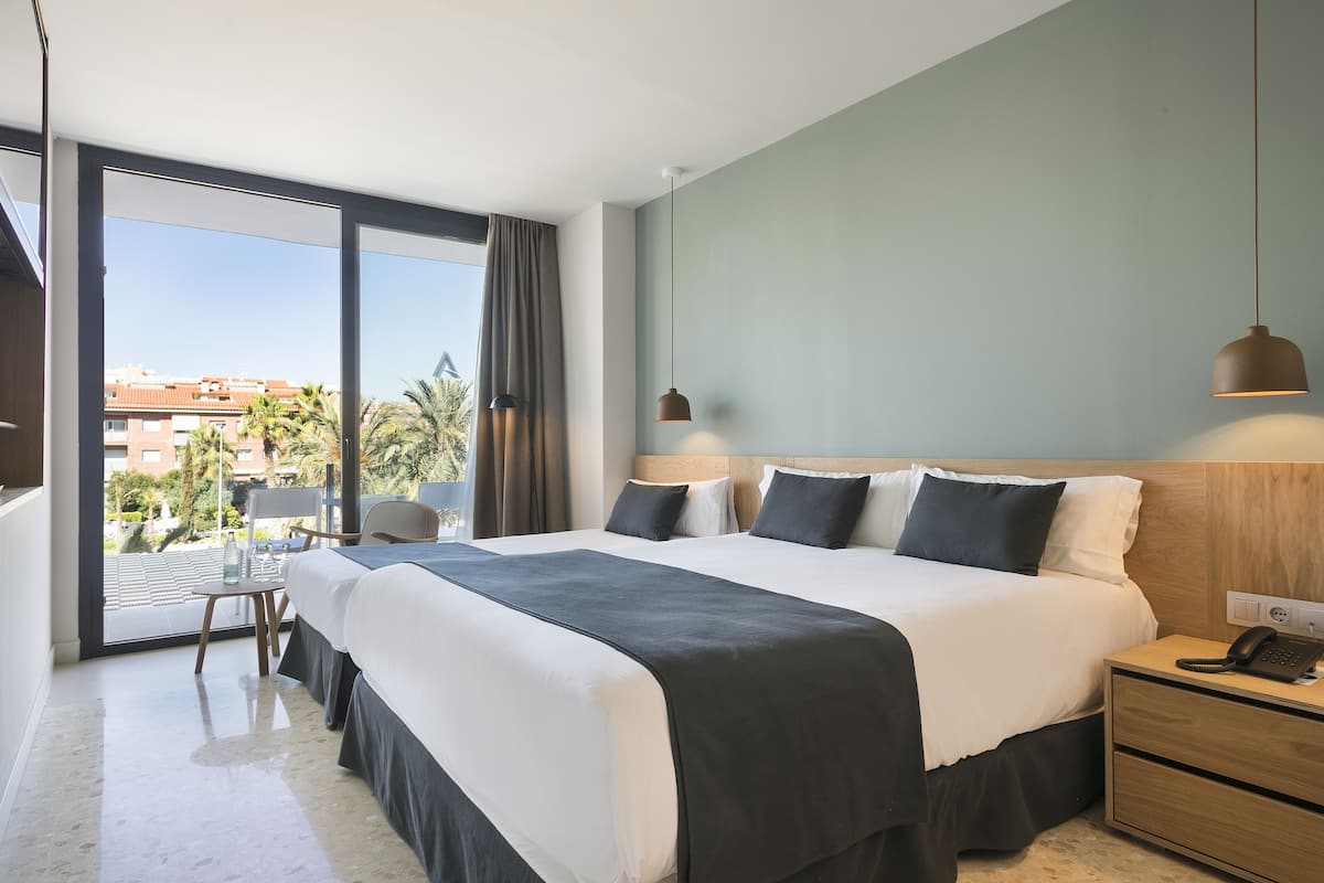 Ein modernes Hotelzimmer mit einem großen Bett, einem Nachttisch mit Telefon und Blick auf die Stadt durch große Fenster.
