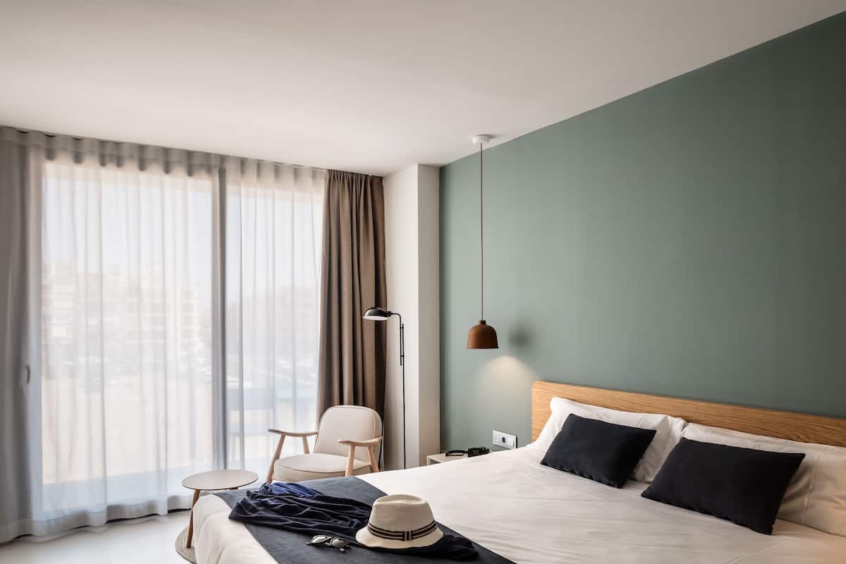 Ein modernes Hotelzimmer mit einem großen Bett, einem kleinen runden Tisch, einem Sessel und Blick auf die Stadt durch transparente Vorhänge.