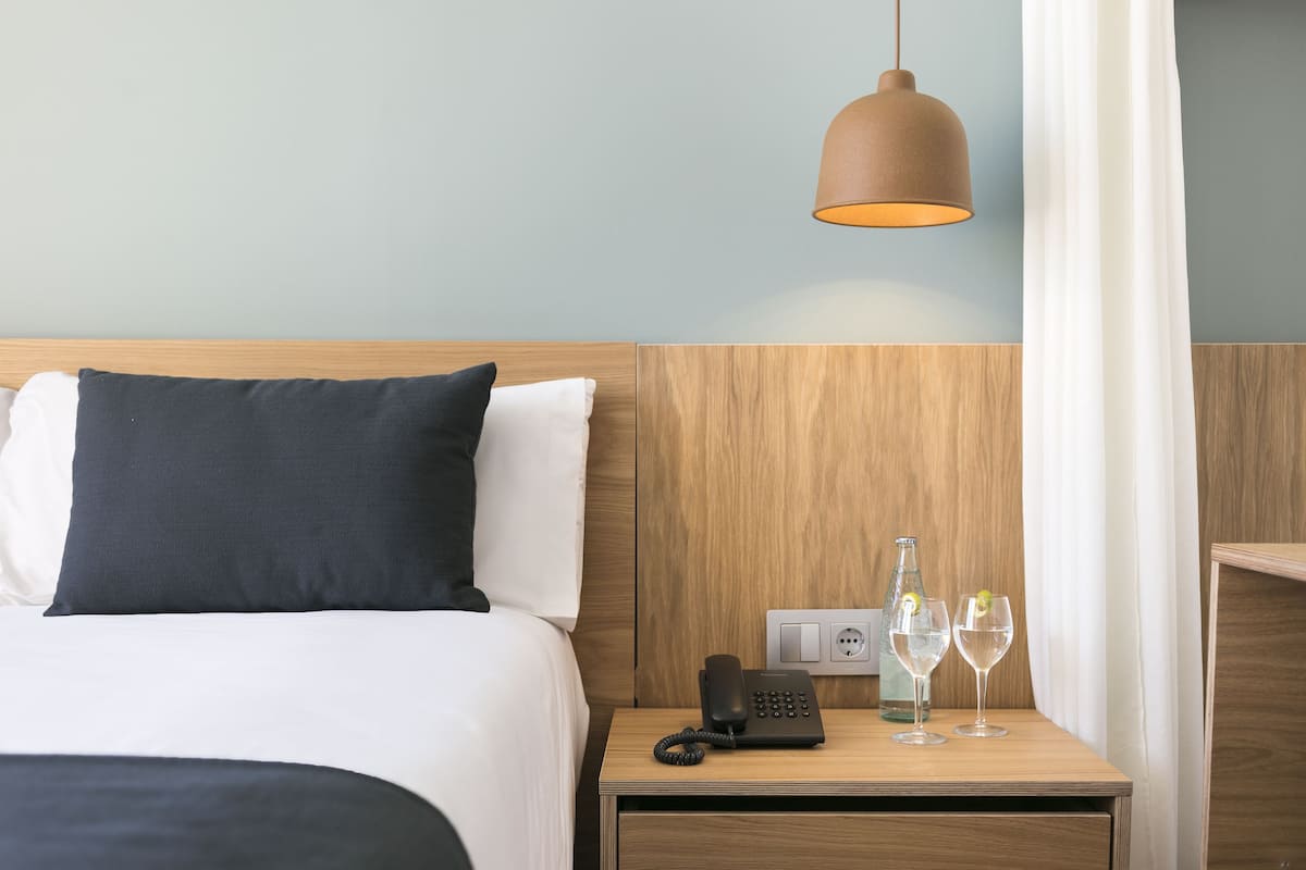 Ein Hotelzimmer mit einem Holzkopfteil, einem Bett mit weiß und schwarz gestreiften Kissen, einem Nachttisch mit Telefon, zwei Gläsern und einer Flasche.