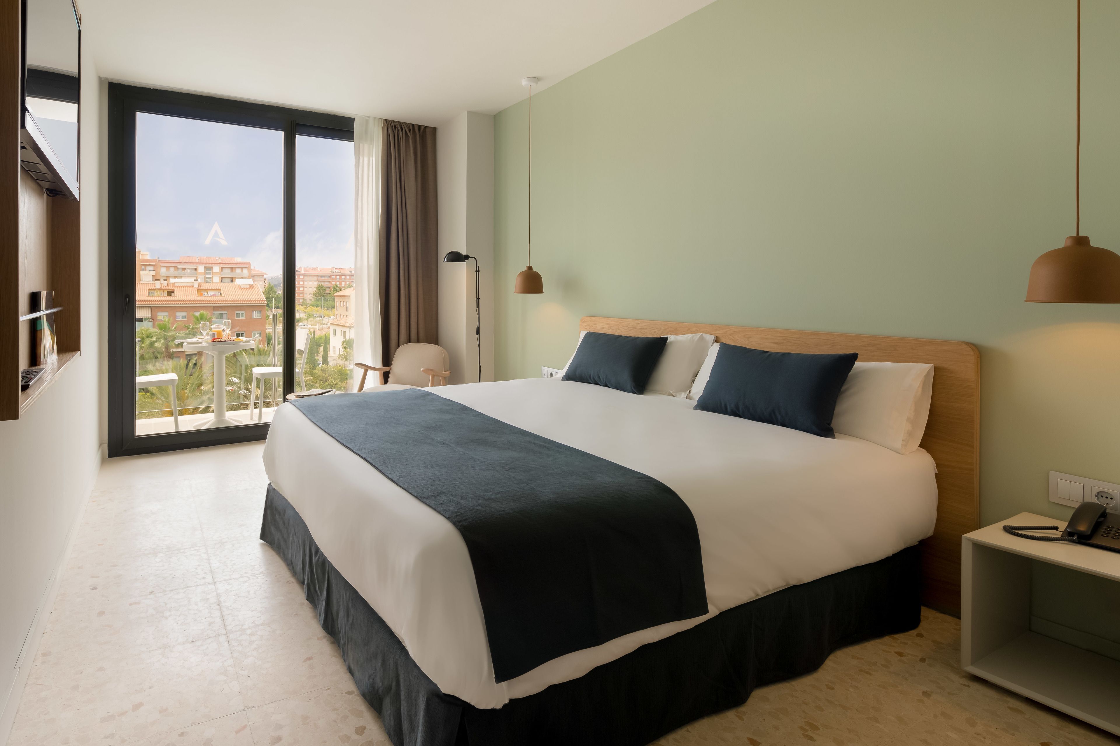 A hotel room with a large bed, a desk, a chair, and a view of the city through a large window.