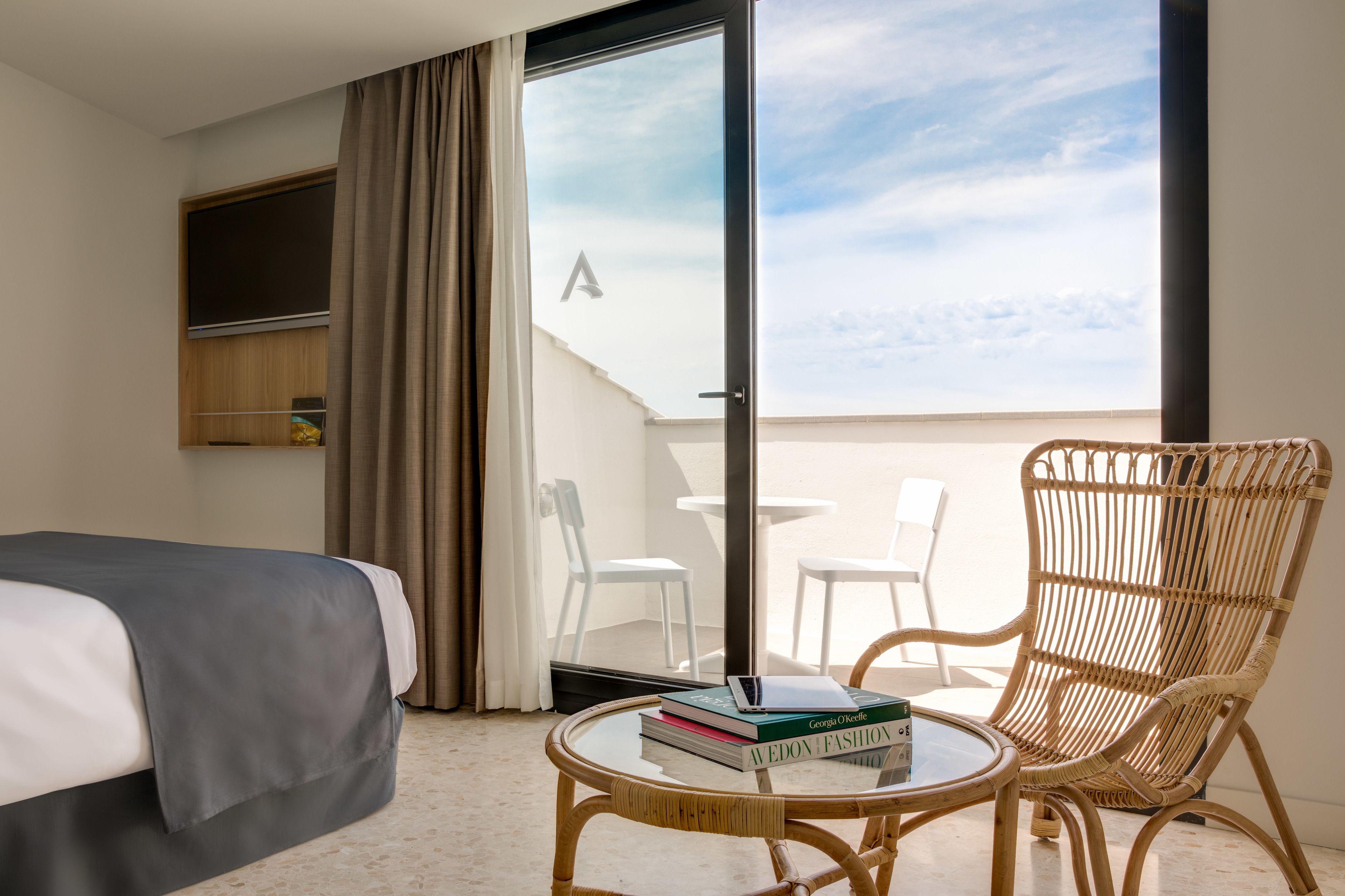 Et hotelværelse med en seng, et rundt bord med bøger, en fletstol og en balkon med hvide stole.
