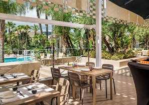 An outdoor dining area with tables and chairs, a pool in the background, and lush greenery.