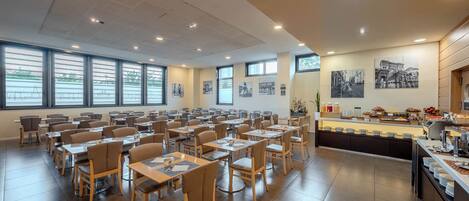 Un comedor amplio con varias mesas y sillas, ventanas grandes y un mostrador de buffet con diferentes alimentos.