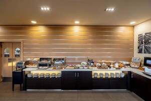 Un'area buffet con diversi cibi e posate, una zona con sedute e una poltrona, e un televisore montato a parete.
