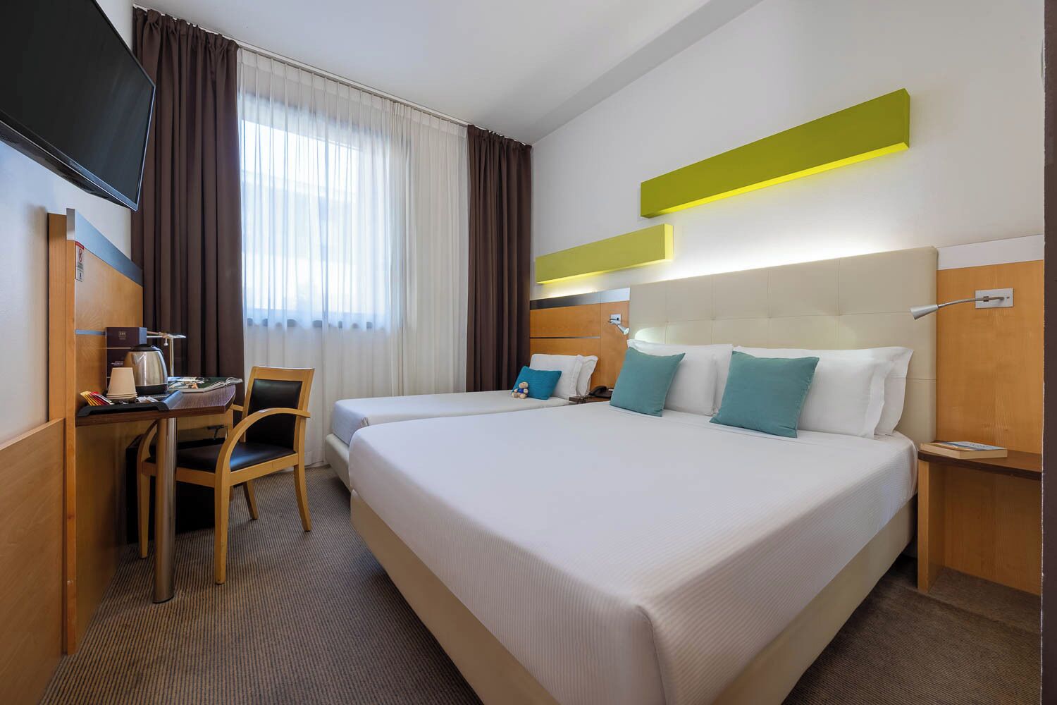 A hotel room with two beds, a desk with a chair, a TV, and a window with curtains.