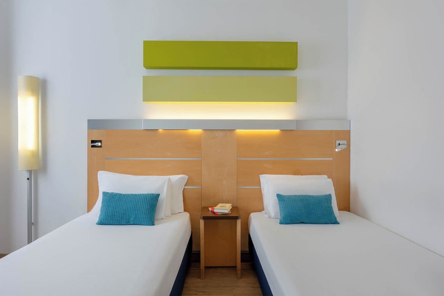 Ein Hotelzimmer mit zwei Einzelbetten, einem Nachttisch, einer Lampe und einem hölzernen Kopfteil.