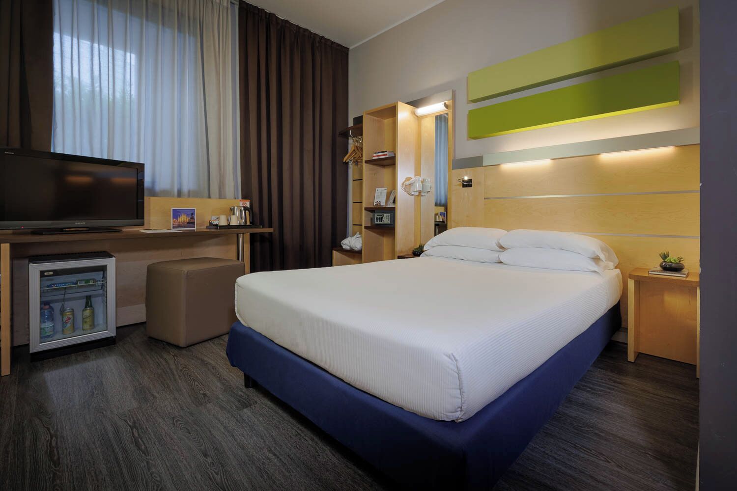 Ein Hotelzimmer mit einem Bett, einem Fernseher, einer Minibar und einem Schreibtisch mit Spiegel.