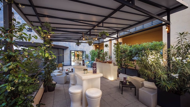 An outdoor seating area with white cushioned chairs and tables, surrounded by potted plants and a bar area. - iH Hotels Milano Gioia (Milan)