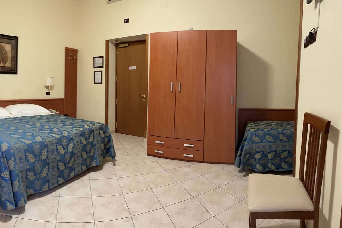 Habitación de hotel con dos camas, un armario de madera, una mesita con un jarrón de flores y una silla.
