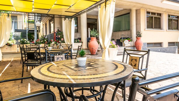 Een buitenterras met ronde tafels en stoelen, potplanten en een geel zonnescherm.
