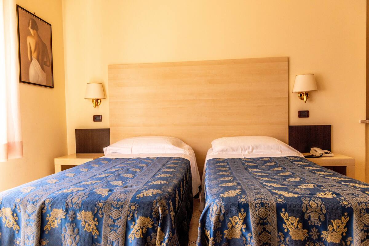 Habitación de hotel con dos camas, cada una con colcha azul estampada de flores, cabeceros de madera, lámparas de noche y mesitas de noche con teléfono.